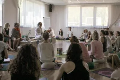A large group of people sitting on the floor in a bright room, listening to a female instructor.