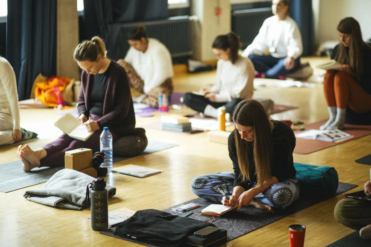 A group of people sitting on yoga mats in a bright studio, writing in journals and notebooks during a workshop.