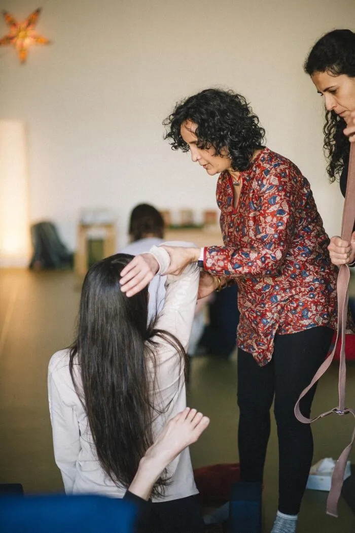 A yoga instructor in a red floral print tunic providing a hands-on adjustment to a student's arm and head during a session.