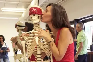 A woman in a red tank top kissing the cheek of a medical skeleton model wearing a red blindfold.