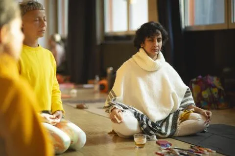 A group of people sitting cross-legged on the floor in a brightly lit room, practicing focused meditation with their eyes closed.