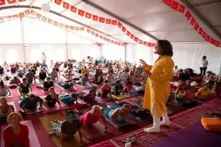 A large group of people practicing yoga in a white tent, led by an instructor in a yellow tunic standing on a stage.