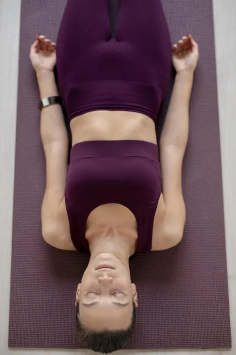 Overhead view of a woman resting in yoga corpse pose on a purple mat.
