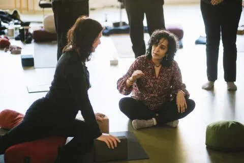A yoga instructor sitting cross-legged on the floor, talking to a student during a class.